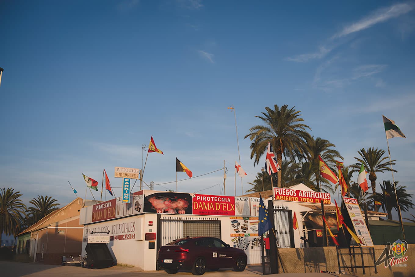 Caseta de pirotecnia de temporada con banderas y rótulos de fuegos artificiales junto a palmeras